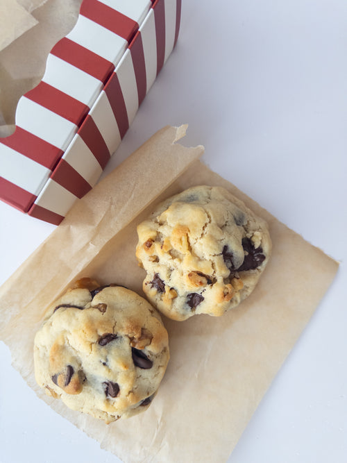 Two thick chocolate chip walnut cookies on brown paper beside a red and white striped gift box