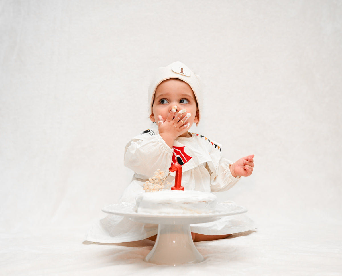 Baby tasting a white first birthday smash cake with a red 1 candle on a cake stand