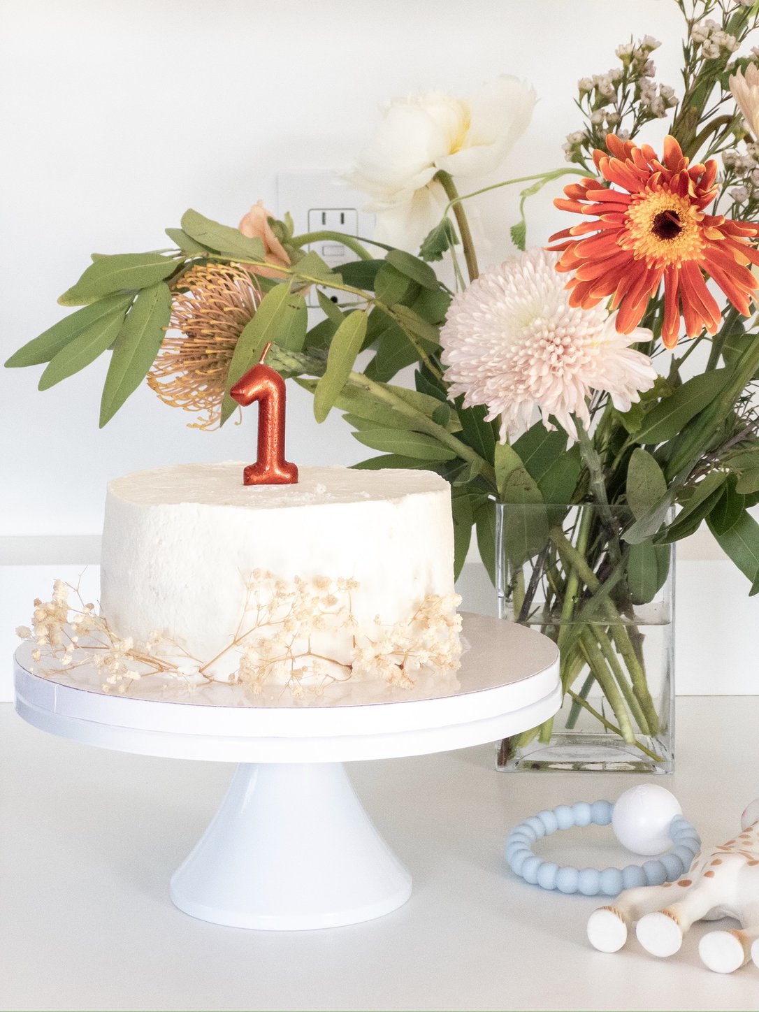 First birthday cake with a copper 1 candle on a white cake stand, with flowers behind
